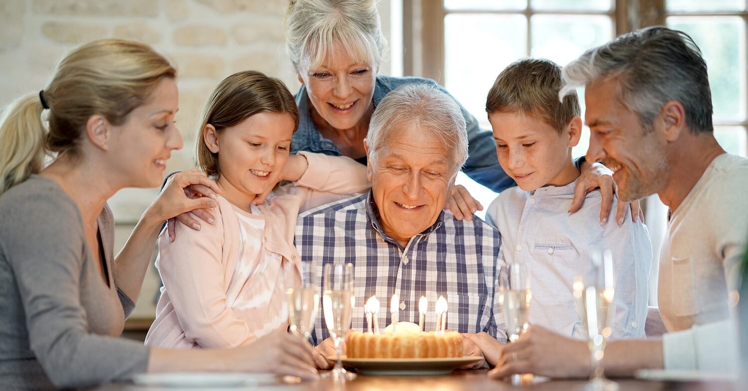 Opa hat Geburtstag und sitzt vor einem Kuchen mit Kerzen, seine Familie steht neben ihm und gratuliert