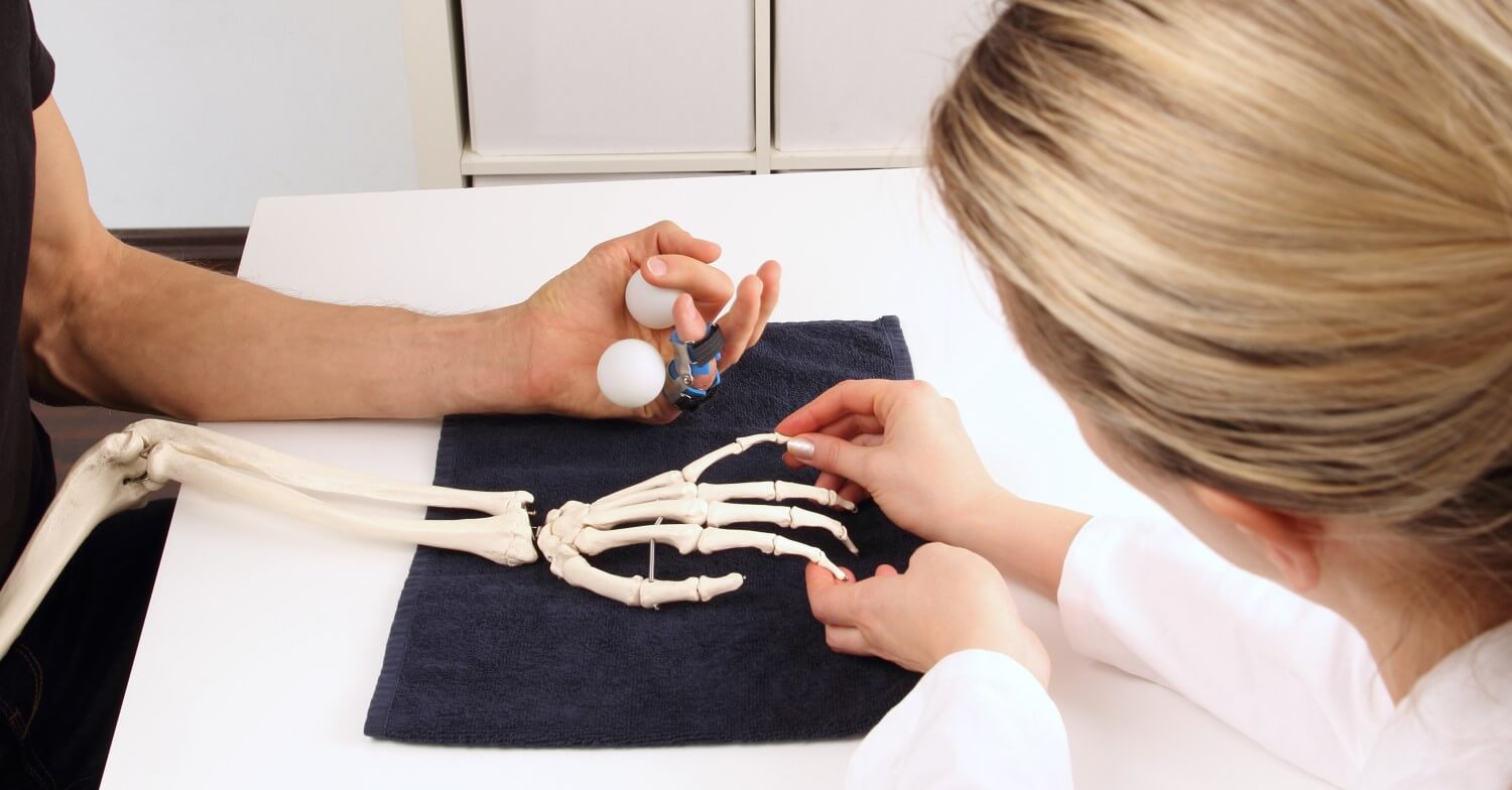 Eine Ärztin erklärt einem Patienten, der eine Schiene am Finger trägt mit Hilfe eines Armskelettes die verschiedenen Knochen der Hand 