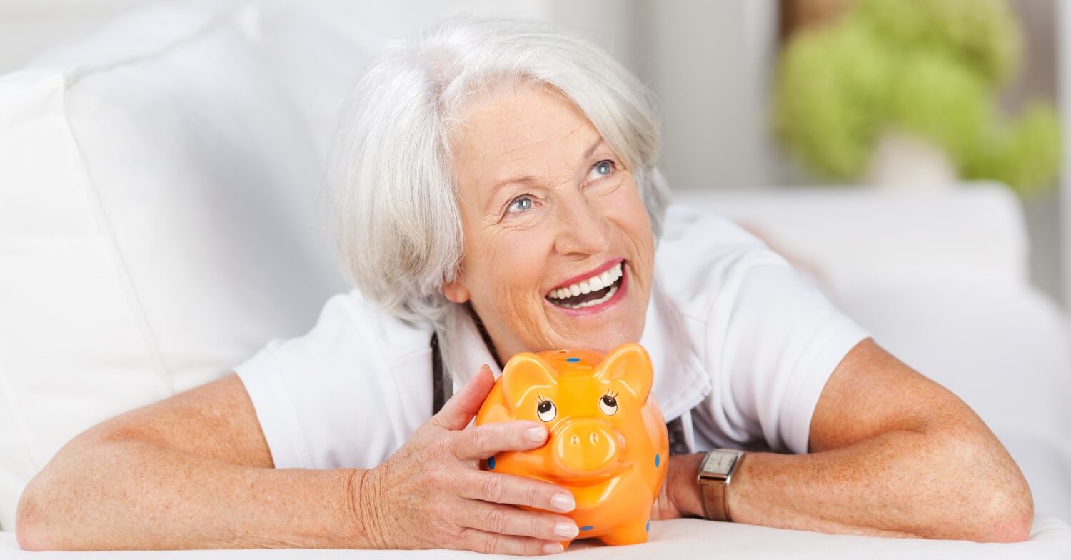 Eine lachende Seniorin mit weißen Haaren hält ein orangenes Sparschwein vor sich