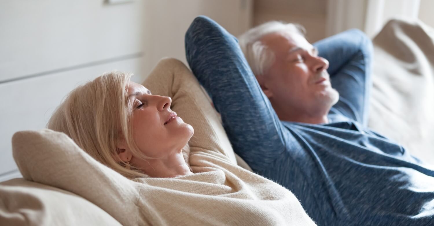 Ein Rentnerpaar sitzt auf dem Sofa. Beide haben die Arme hinter dem Kopf verschränkt und sich zurückgelehnt