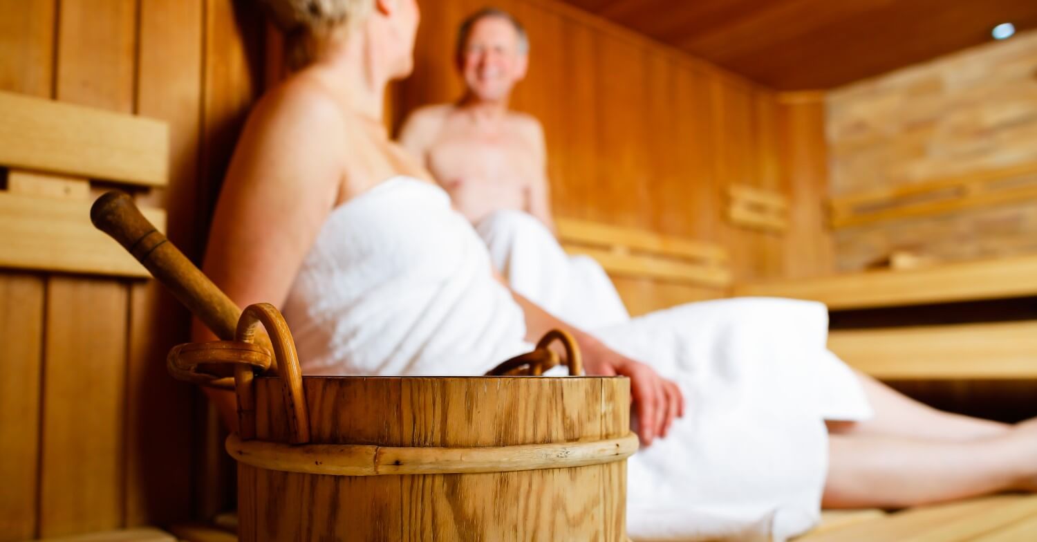 Ein Rentnerpaar sitzt in weiße Handtücher gehüllt in einer finnischen Sauna, im Vordergrund steht ein Holzeimer für den Aufguss
