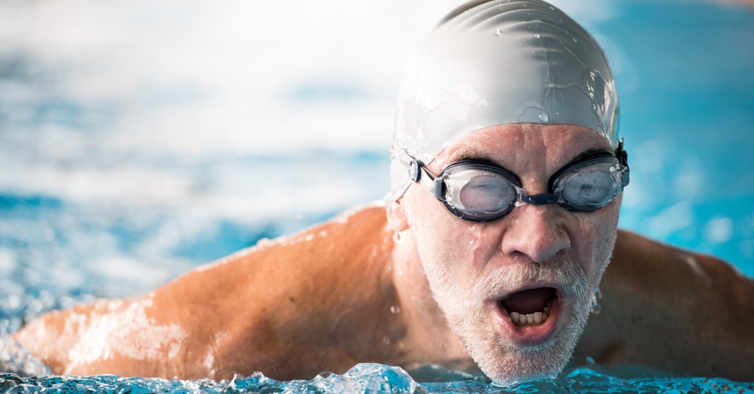 Ein älterer Mann mit grauem Bart schwimmt im Schwimmbad. Er trägt eine Badekappe und eine Schwimmbrille.
