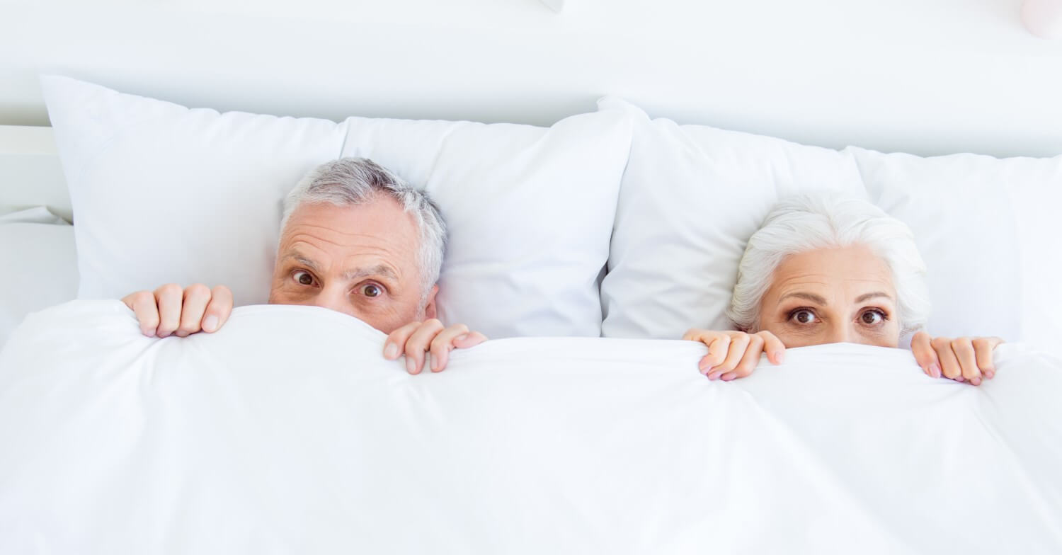 Ein Seniorenpaar liegt im Bett. Beide haben die Bettdecke bis zur Nasenspitze hochgezogen.