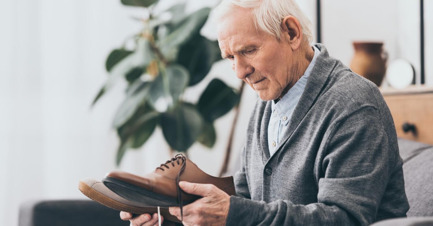 Ein grauhaariger Senior hält seine beiden Schuhe in der Hand und schaut sie fragend an