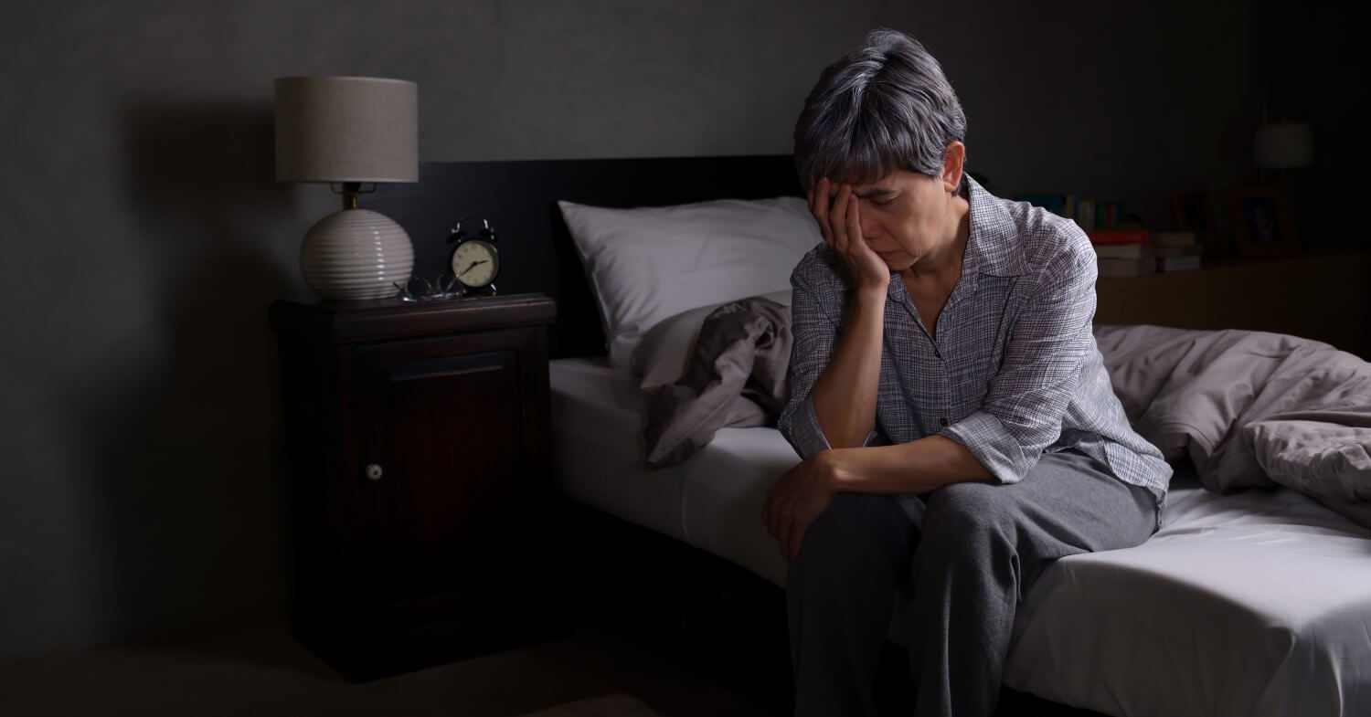 Depressive Seniorin Eine Seniorin mit kurzen grauen Haaren sitzt auf der Bettkante und hat den Kopf in die Hand gestützt. Sie wirkt traurig.