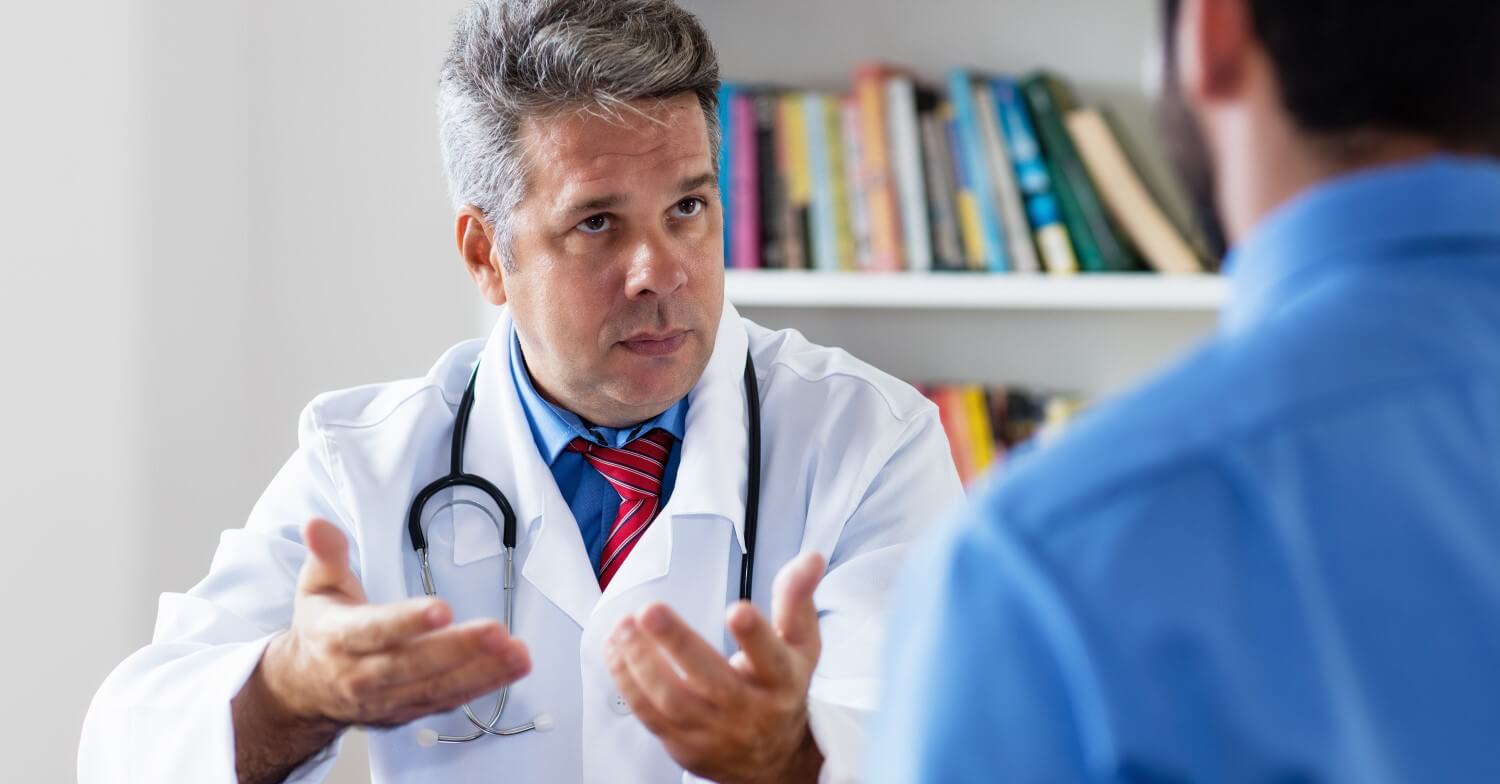 Ein Arzt im weißen Kittel hat sein Stetoskop um den Nacken gelegt, erklärt seinem Patienten etwas. Vom Patient ist nur der Rücken zu sehen.