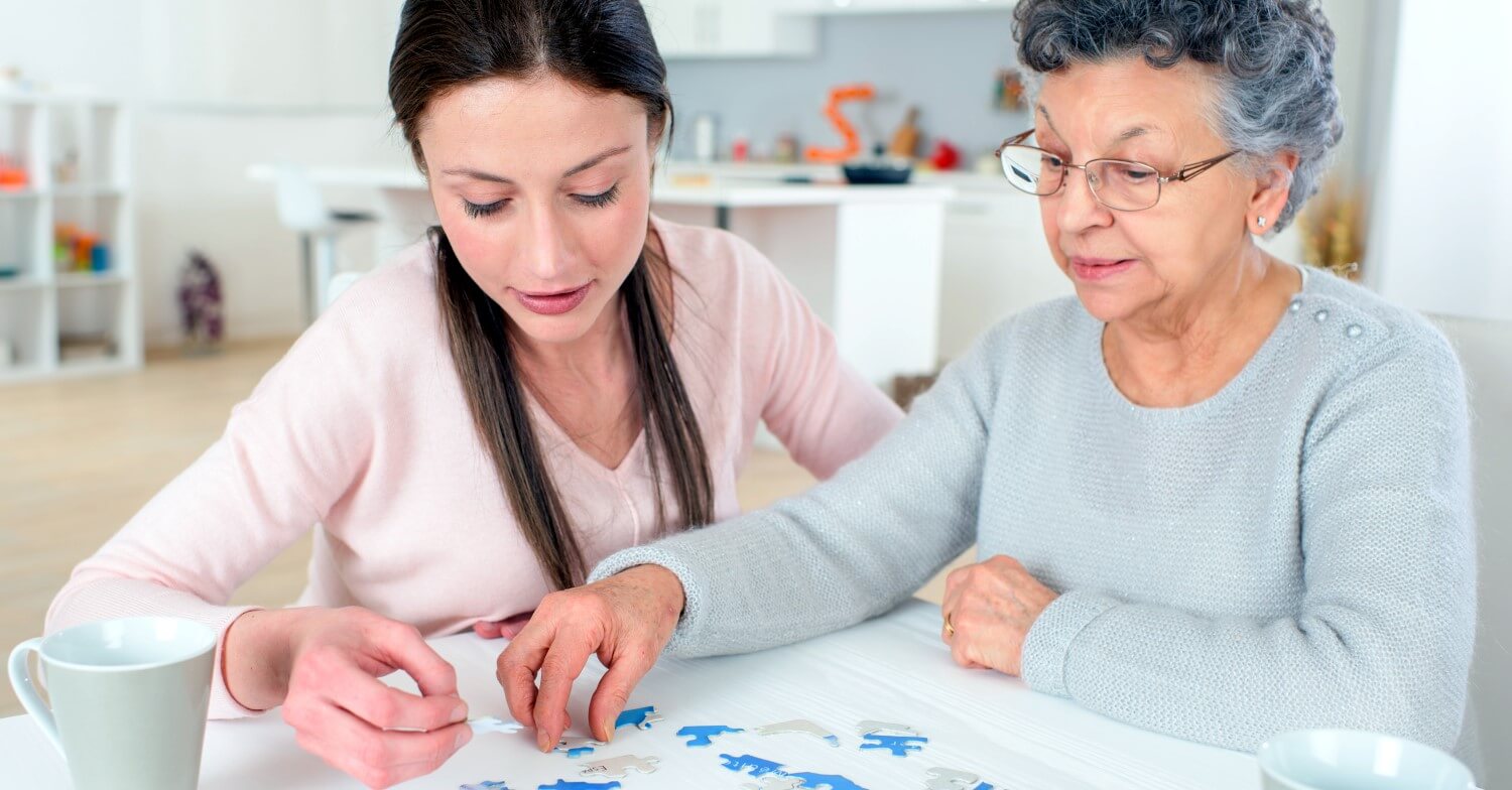 Eine Seniorin sitzt am Tisch und versucht ein Puzzle zu machen, eine Pflegerin sitzt neben ihr und hilft ihr