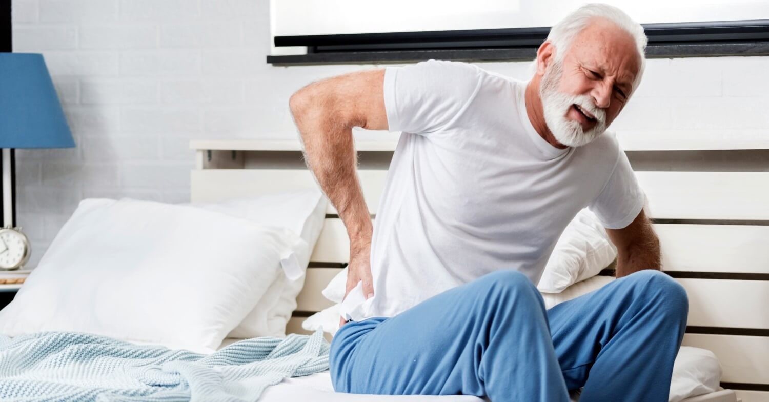 Ein weißhaariger Senior sitzt im Schlafanzug auf der Bettkante und hält sich mit schmerzverzerrtem Gesicht den Rücken