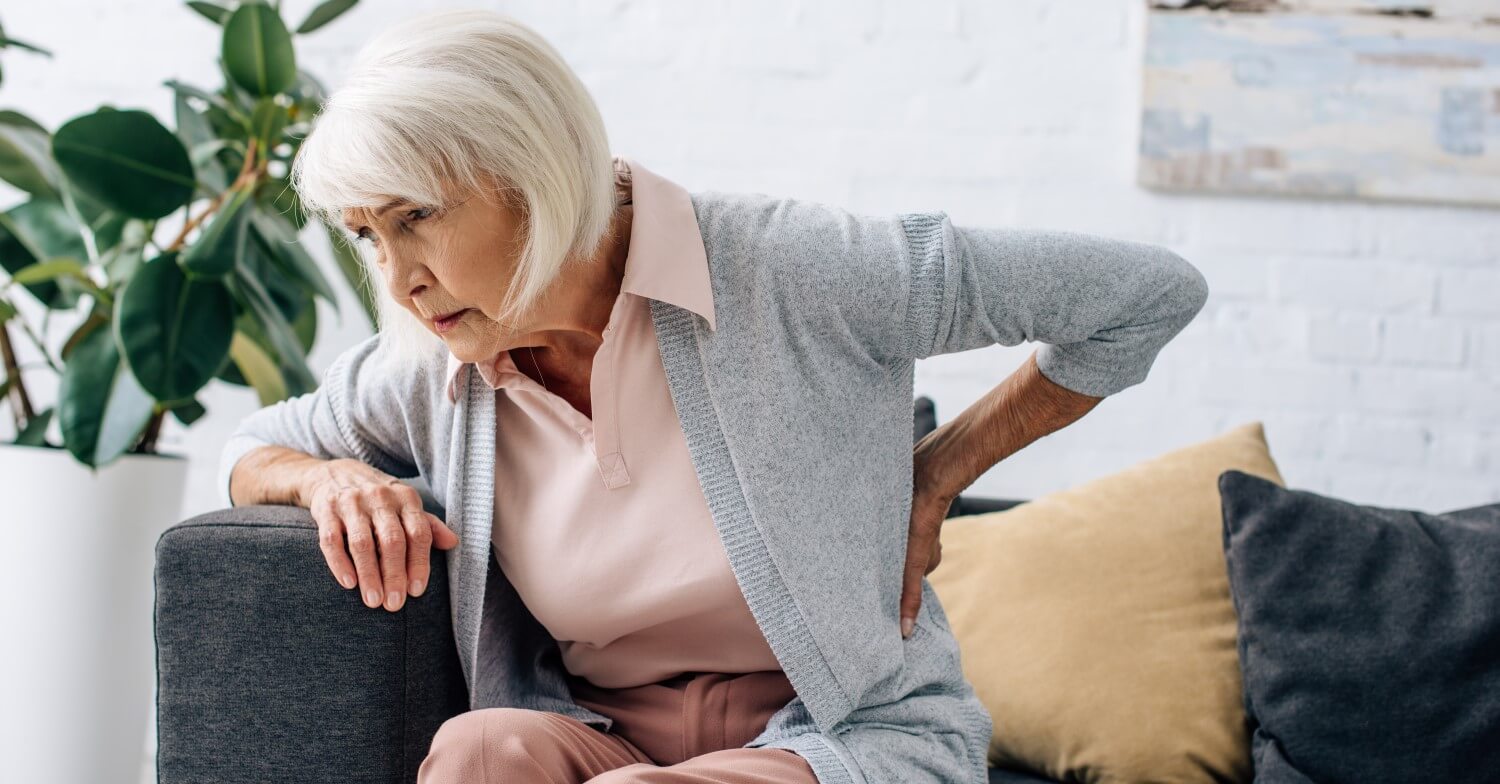 Eine Seniorin sitzt vorgebeugt auf dem Sofa, sie hält sich den Rücken und hat ein schmerzverzerrtes Gesicht