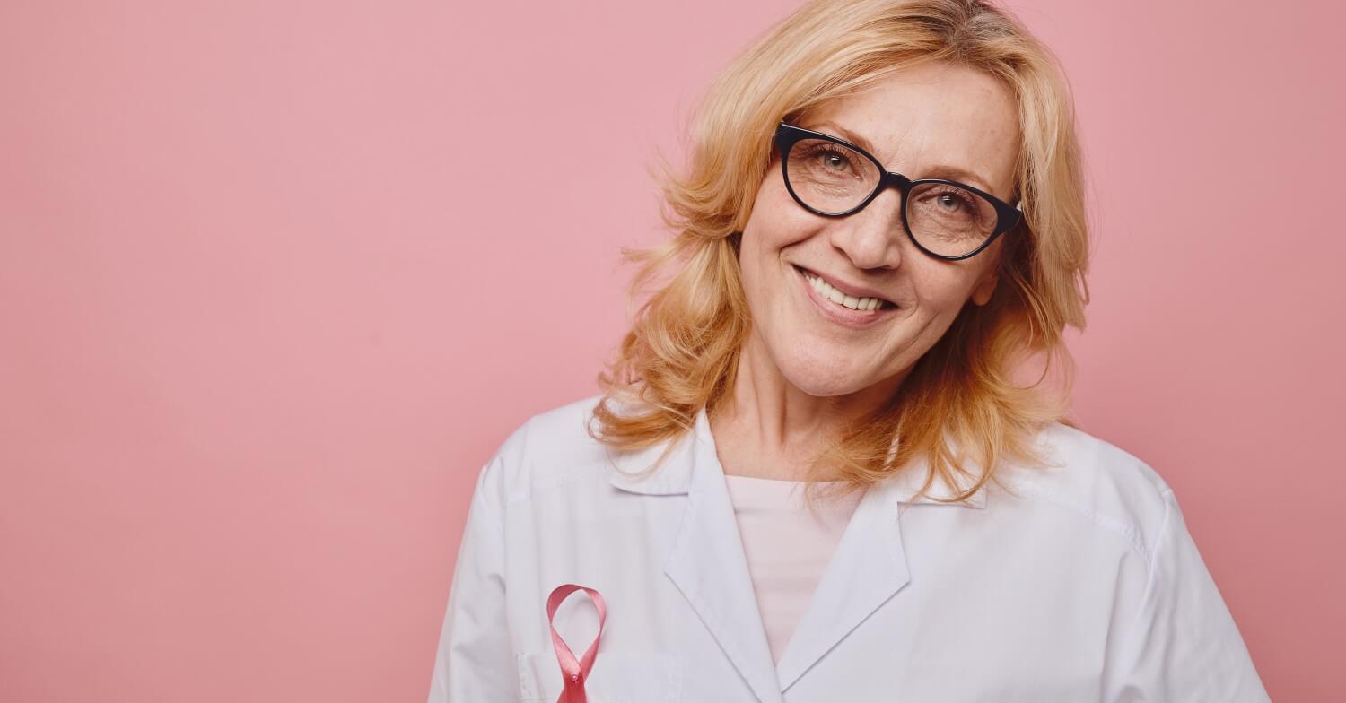 Eine Frau mit weißem Arztkittel blickt in die Kamera, sie hat die Rosa Schleife als Symbol gegen Brustkrebs angehftet