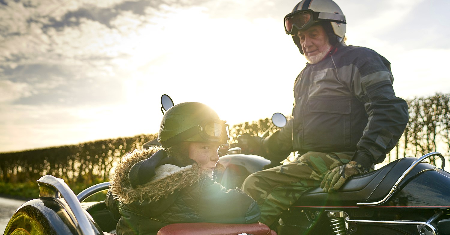 Ein Junge und sein Großvater sitzen auf einem Motorrad mit Beiwagen. Sie haben in Weinbergen angehalten und schauen in die Kamera. Beide tragen Helme.
