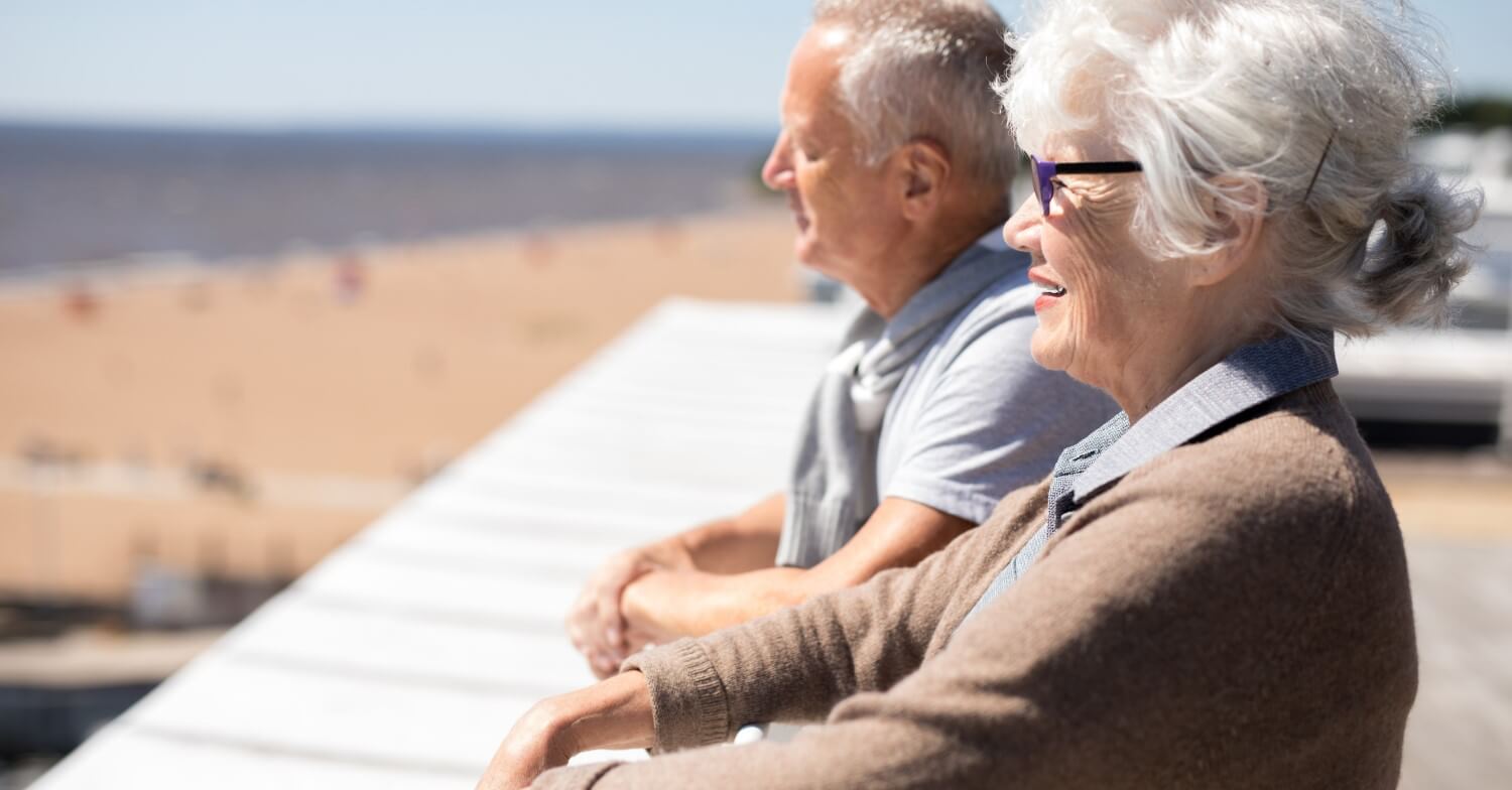 Ein Seniorenpaar lehnt an einem Geländer und schaut auf den Strand