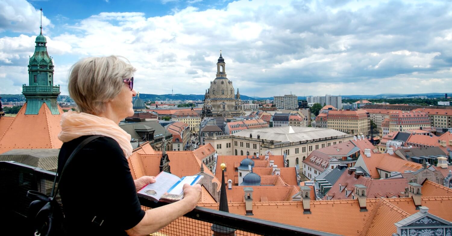 Eine Seniorin steht auf einer Terrasse und blickt auf die Dächer Dresdens