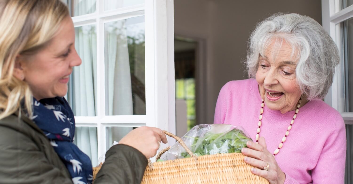 Eine jüngere Frau bringt eine Tasche voller Einkäufe zu einer Seniorin nach Hause