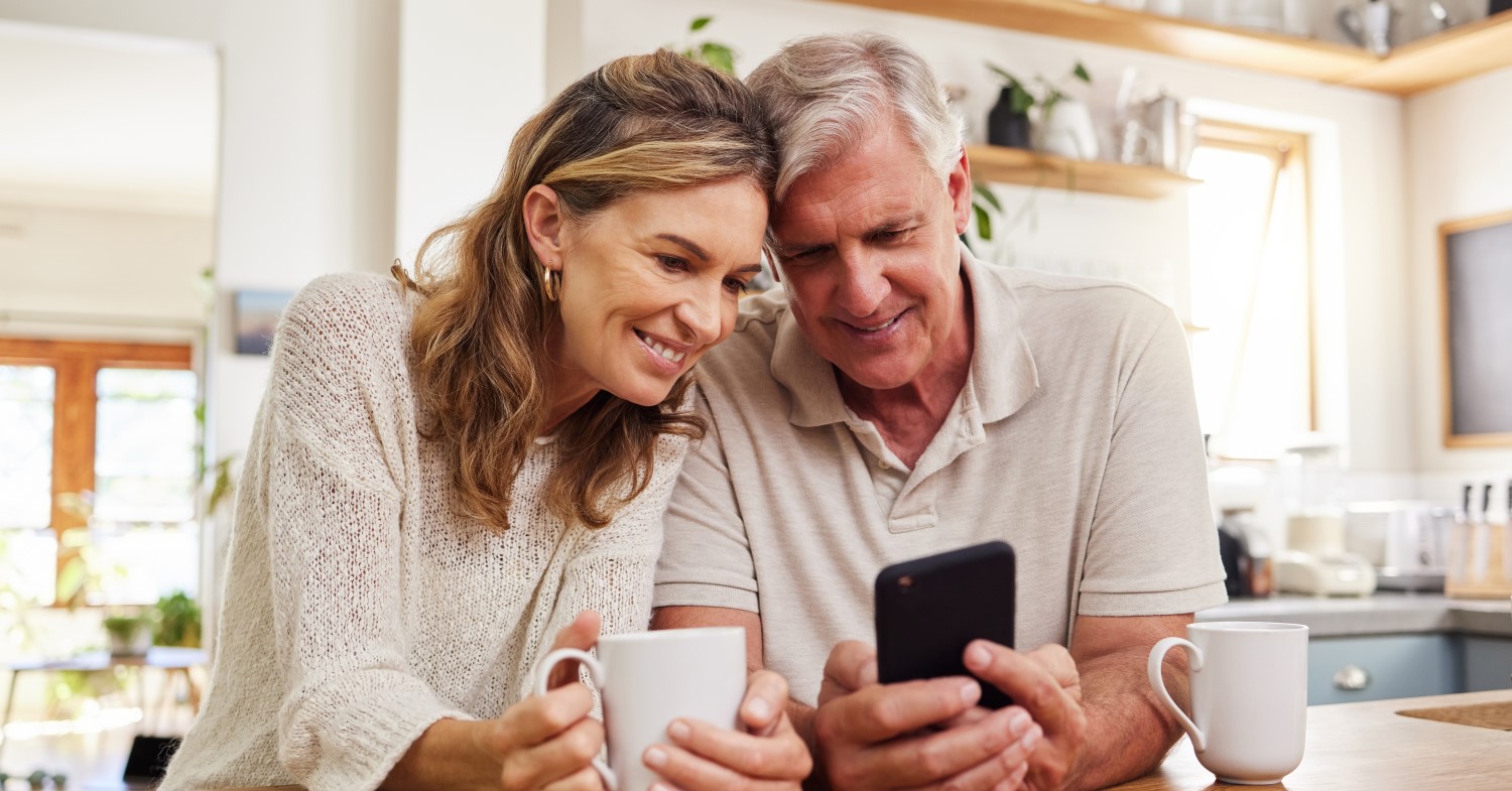Ein älteres Paar sitzt am Tisch und schaut gemeinsam auf ein Smartphone. Beide haben Kaffeetassen vor sich stehen und scheinen glücklich und entspannt zu sein.