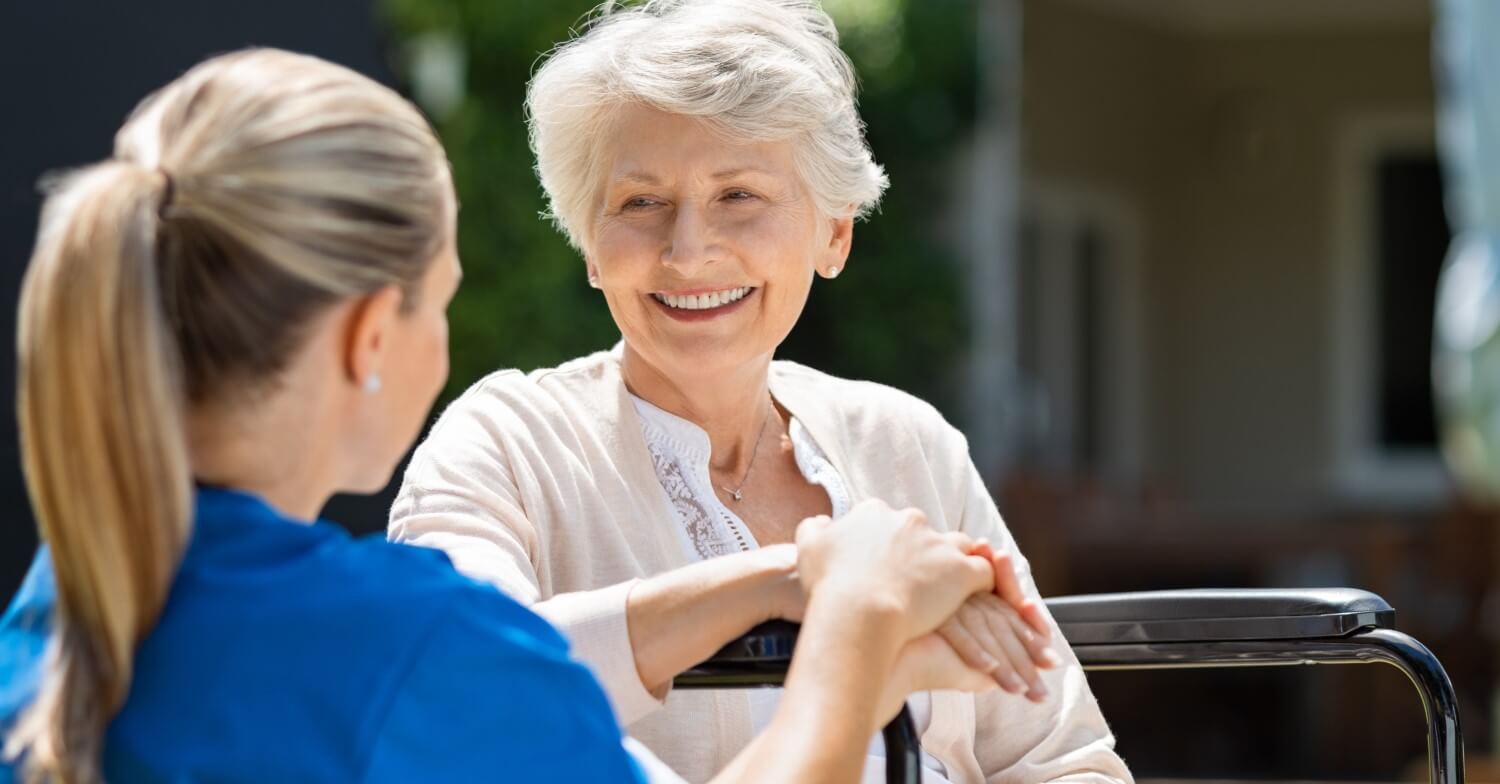 Eine Pflegerin in blauem Kittel kümmert sich um eine Seniorin, die im Rollstuhl sitzt